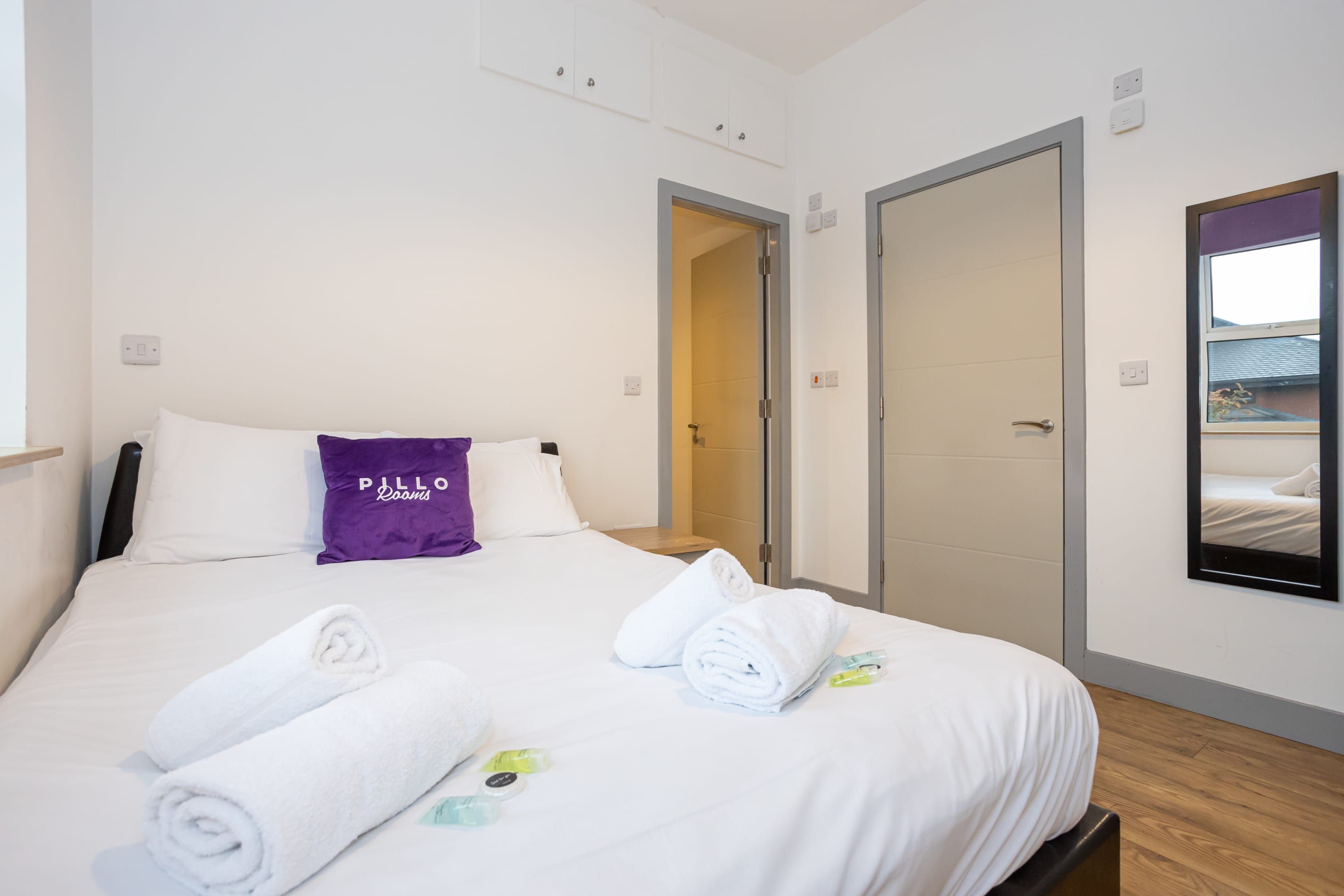 Una habitación de hotel con una cama, ropa de cama blanca, un almohadón púrpura y un espejo en la pared.
