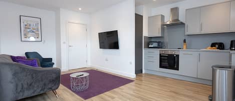 A modern apartment with a grey sofa, a purple rug, a kitchen with grey cabinets, and a flat-screen TV mounted on the wall.