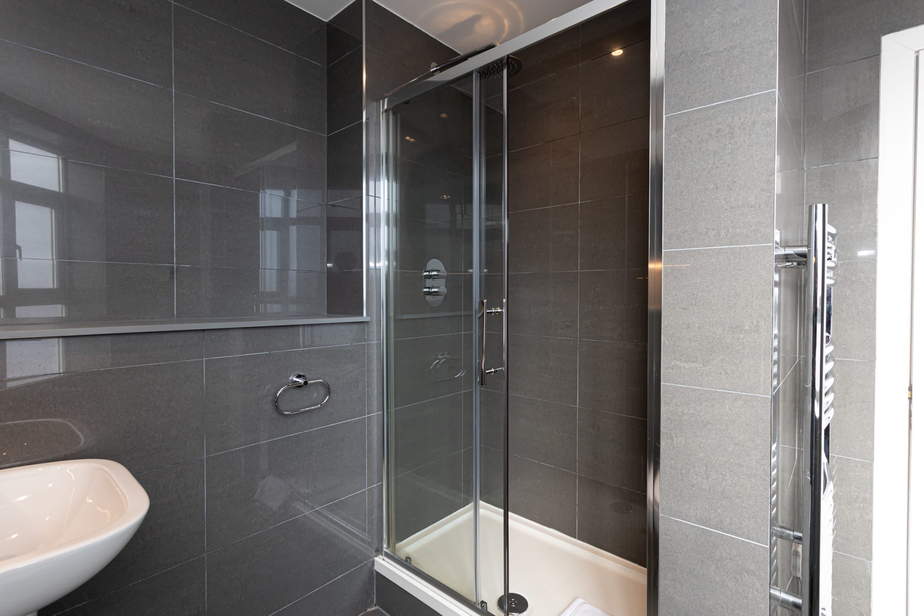 A modern bathroom with a glass-enclosed shower, a white sink, and dark tiled walls.