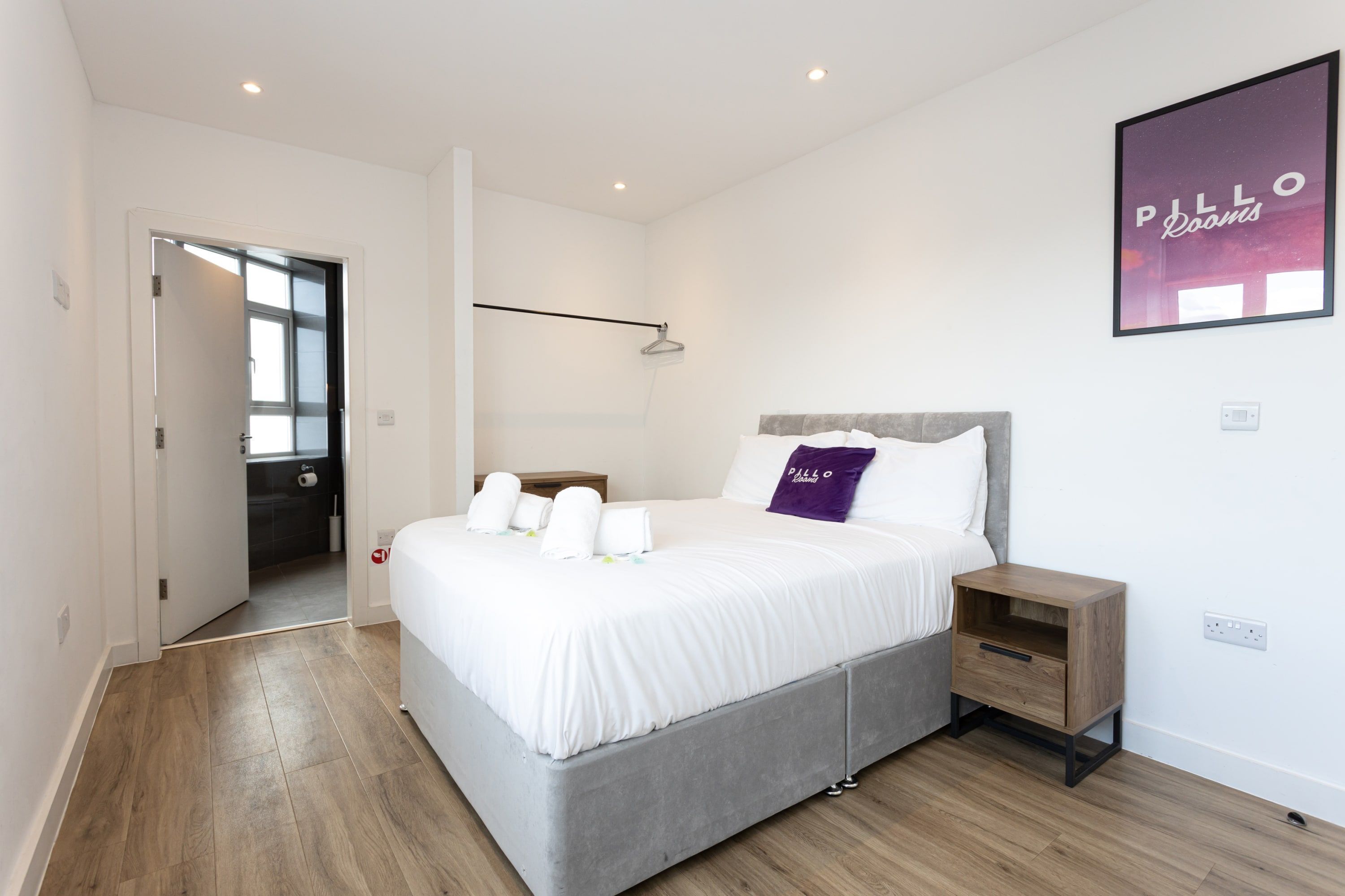 A modern bedroom with a large bed, a nightstand, a wall-mounted shelf, and a door leading to a bathroom.