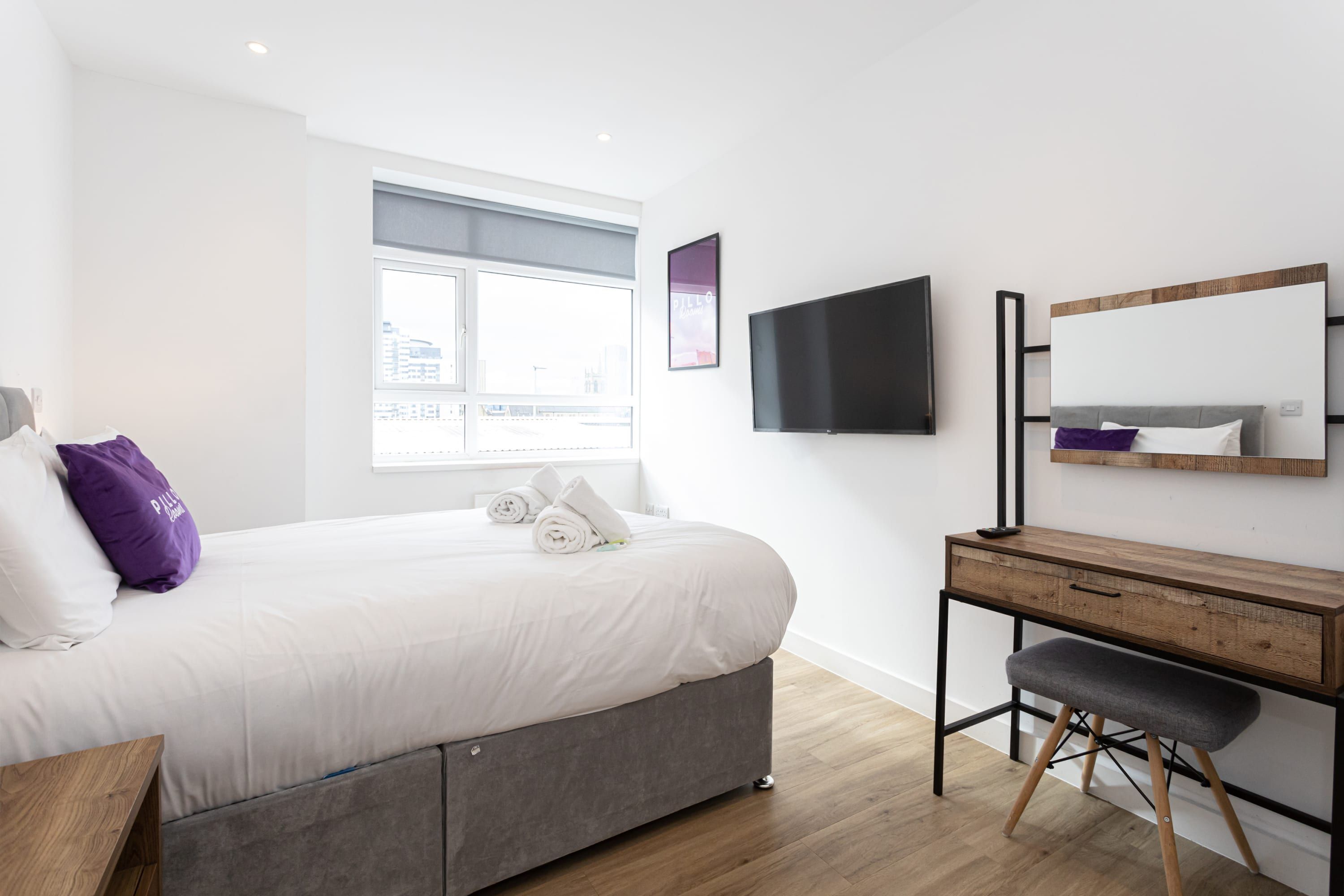 A modern hotel room with a bed, a desk, a chair, a flat-screen TV, and a window with a view of the city.