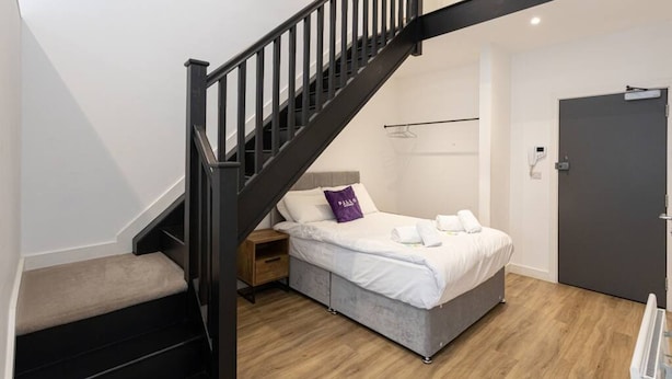 A bedroom with a bed, a nightstand, a dresser, a wooden floor, a staircase, and a door. - Modern and Spacious Duplex in Manchester City Centre (Manchester)