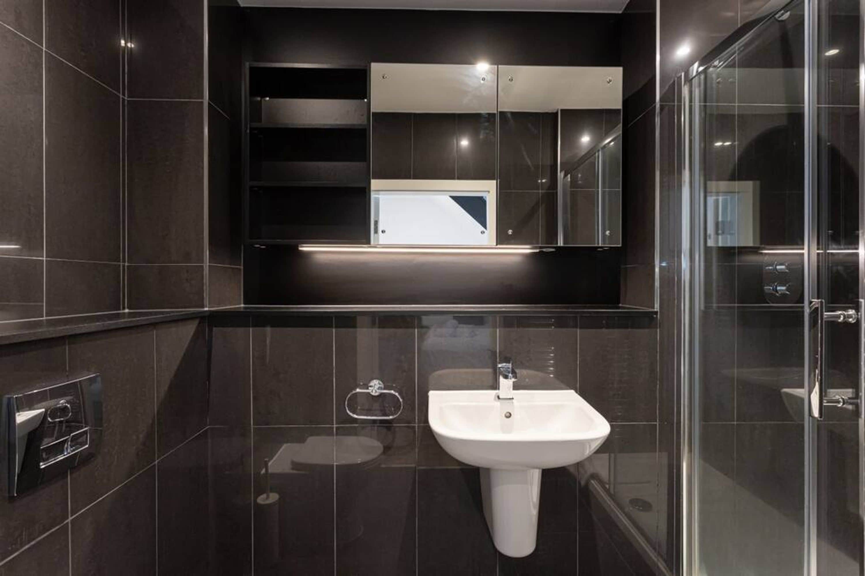 Une salle de bain moderne avec un lavabo blanc, des carreaux foncés, une douche en verre et un meuble-miroir encastré.
