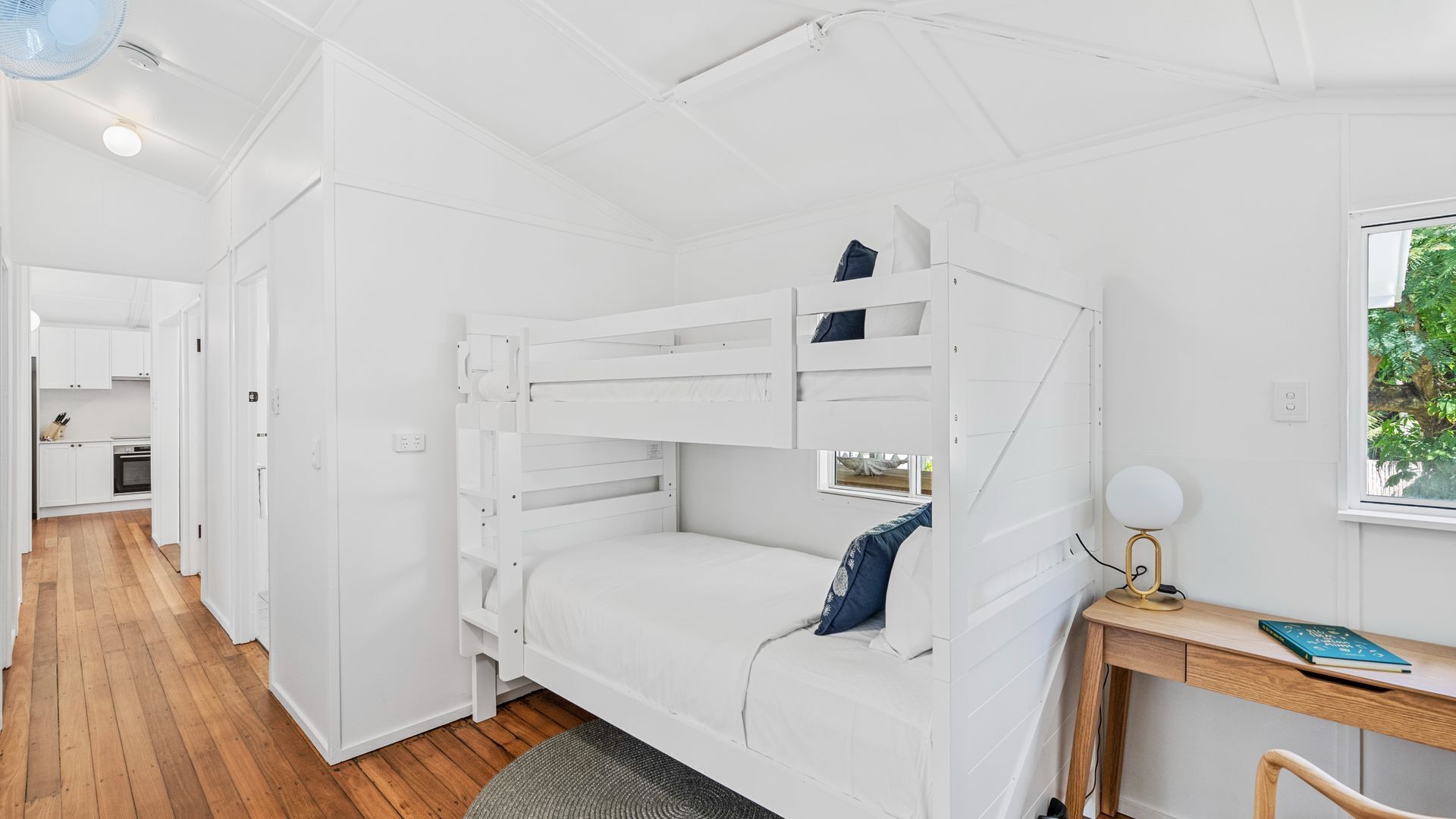 Une chambre avec des lits superposés, un bureau et une chaise, une fenêtre donnant sur un espace verdoyant, et un coin cuisine à l’arrière-plan.