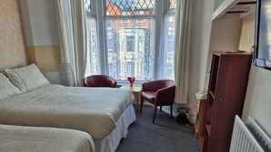 A hotel room with two beds, a small table, two chairs, a radiator, and a window with a view of buildings. - Glendor Hotel (Blackpool)