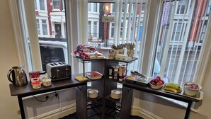 A breakfast setup with a toaster, coffee pot, and various food items on a table near a window with a view of buildings. - Glendor Hotel (Blackpool)
