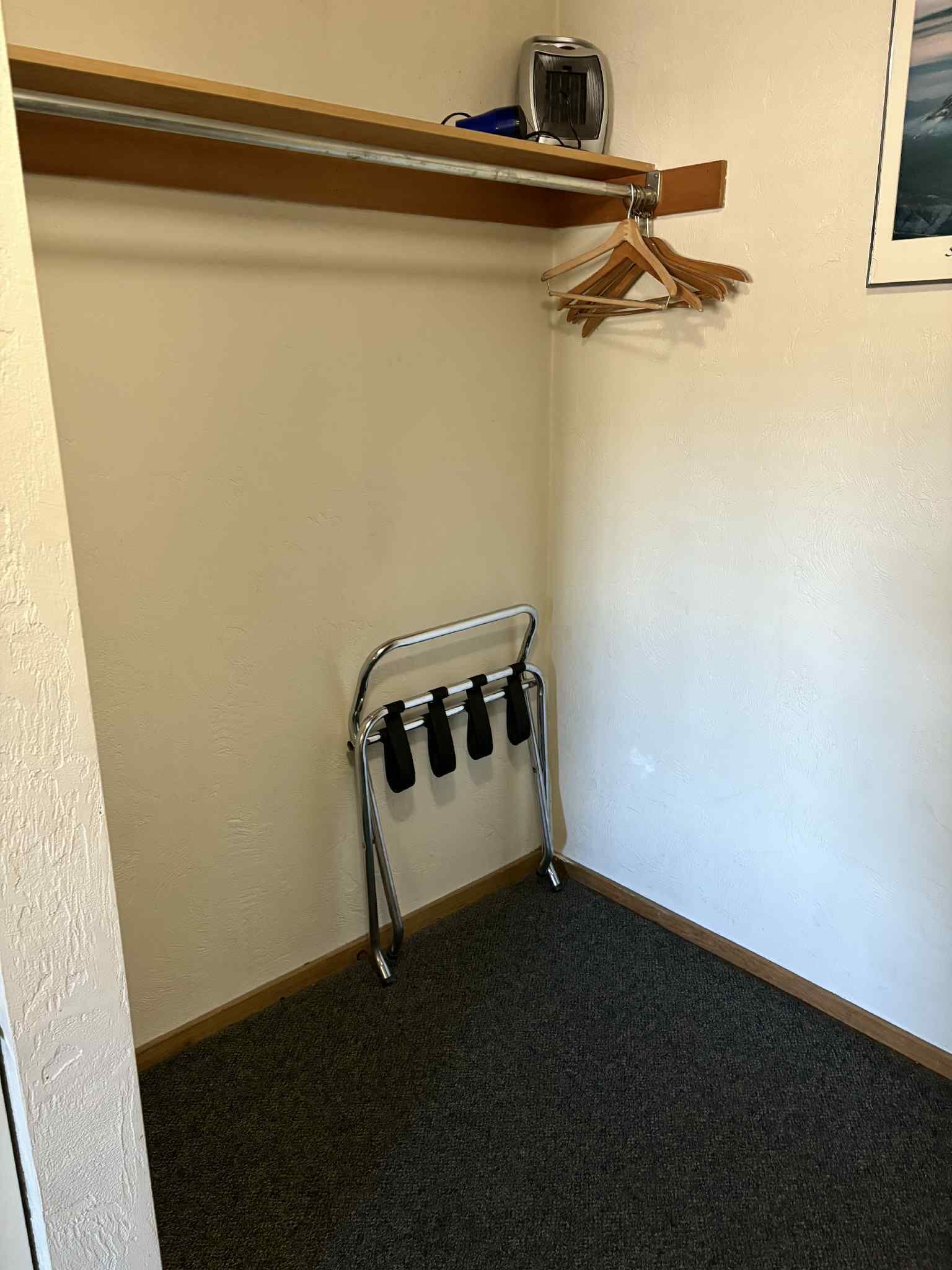 Una esquina de una habitación con un perchero metálico que sostiene cuatro objetos negros, un estante de madera con un objeto azul y un cuadro en la pared.