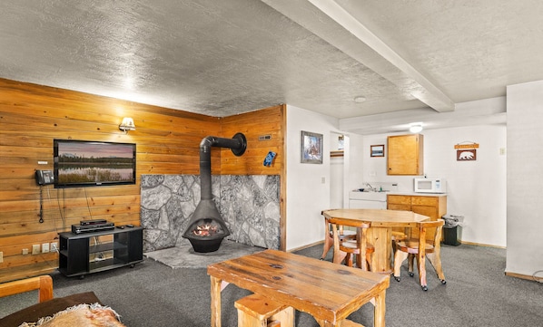 A cozy room with a stone fireplace, wooden walls, a dining table, and a kitchen area. - Willamette Pass Inn (Crescent)