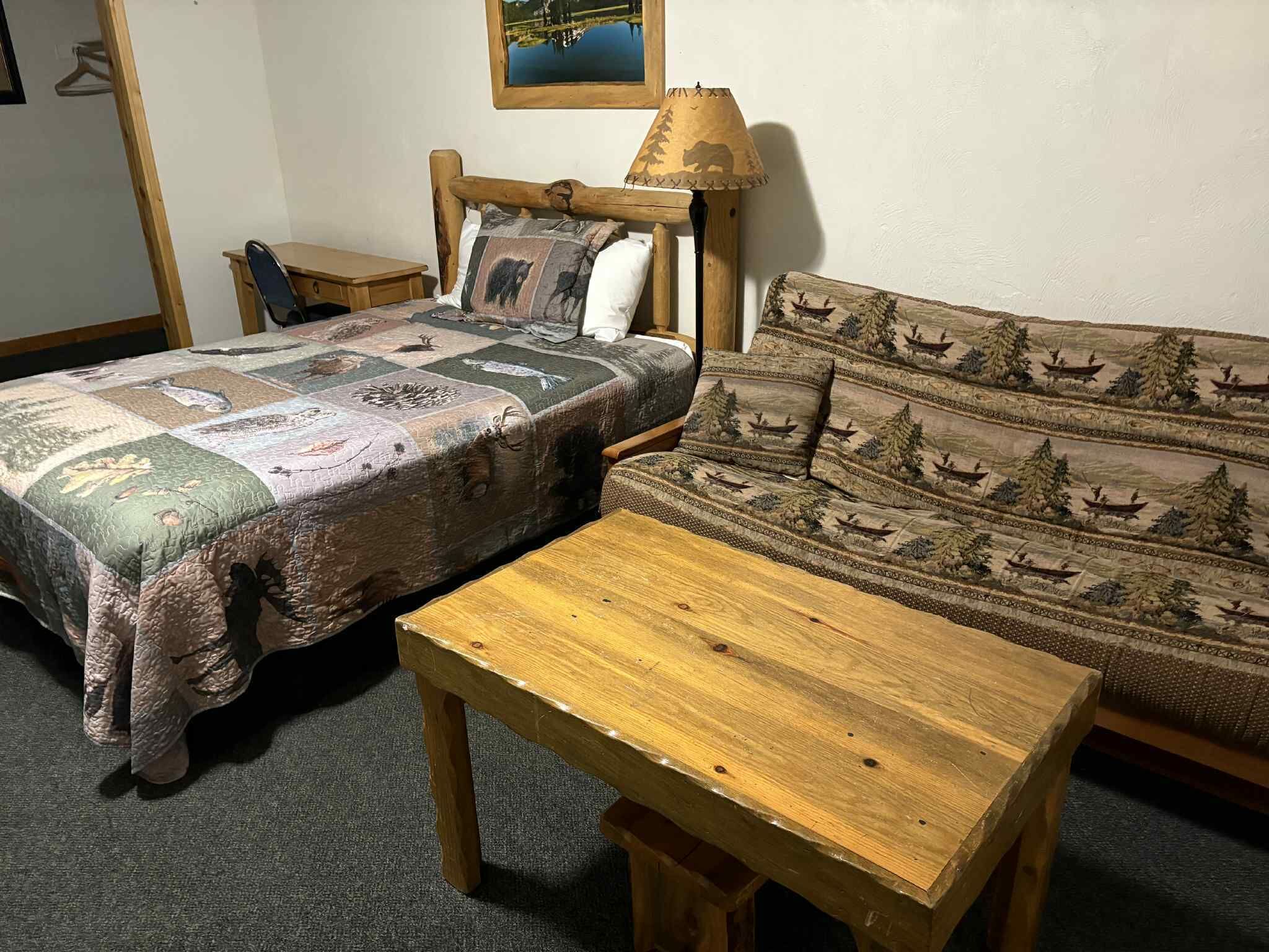 Una habitación con una cama, una mesita de noche de madera, una mesa de madera y un sofá con un cubrecolchón con motivos forestales.