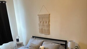 Un dormitorio con cama, mesita de noche, lámpara, un colgador de macramé y una manta amarilla.