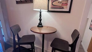 Una habitación pequeña con una mesa redonda de madera y dos sillas, una lámpara y un cuadro en la pared.