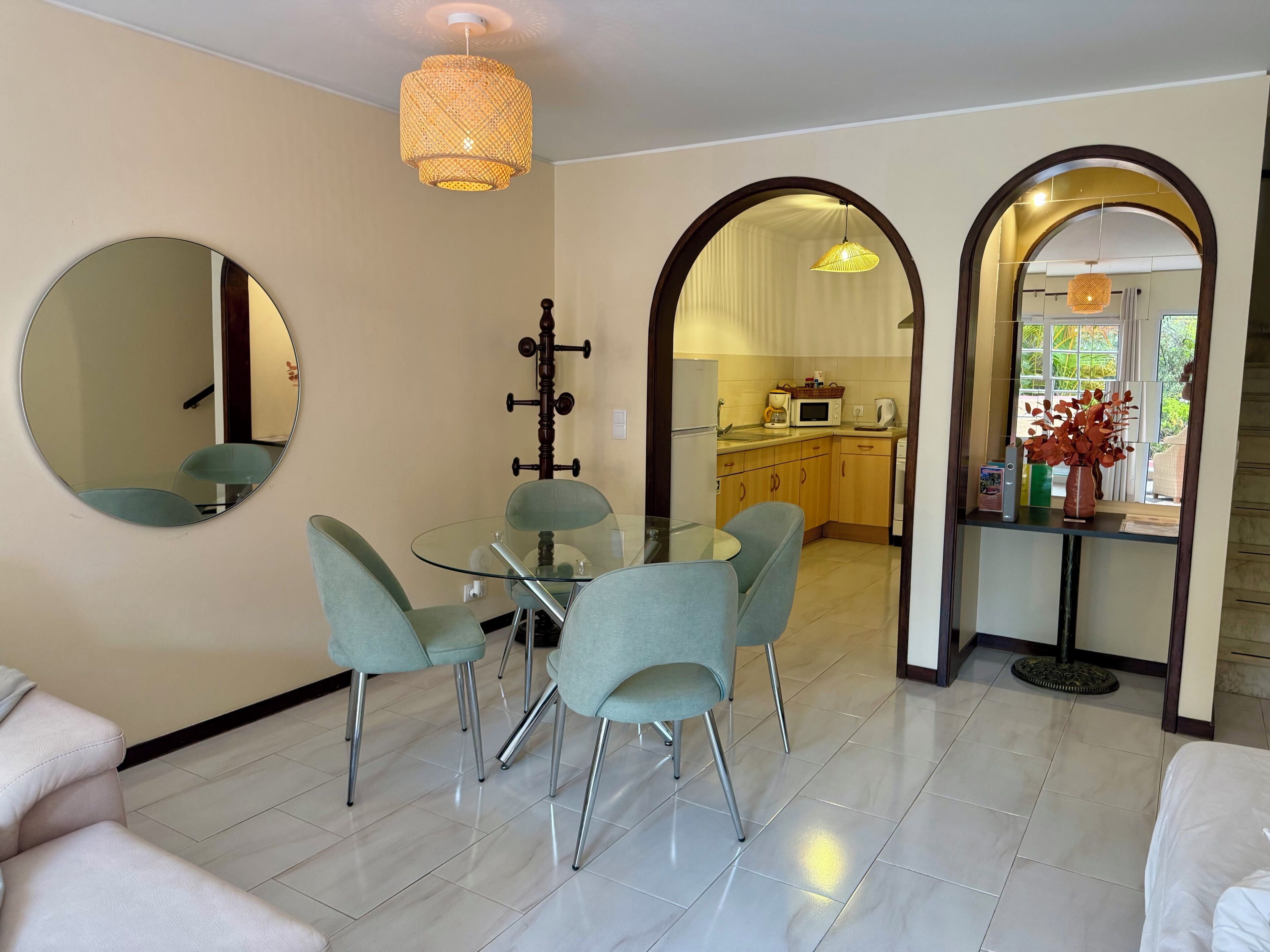 Uma sala de estar moderna com uma mesa de jantar de vidro, cadeiras azul-claras, um espelho redondo e uma cozinha visível através de um arco.