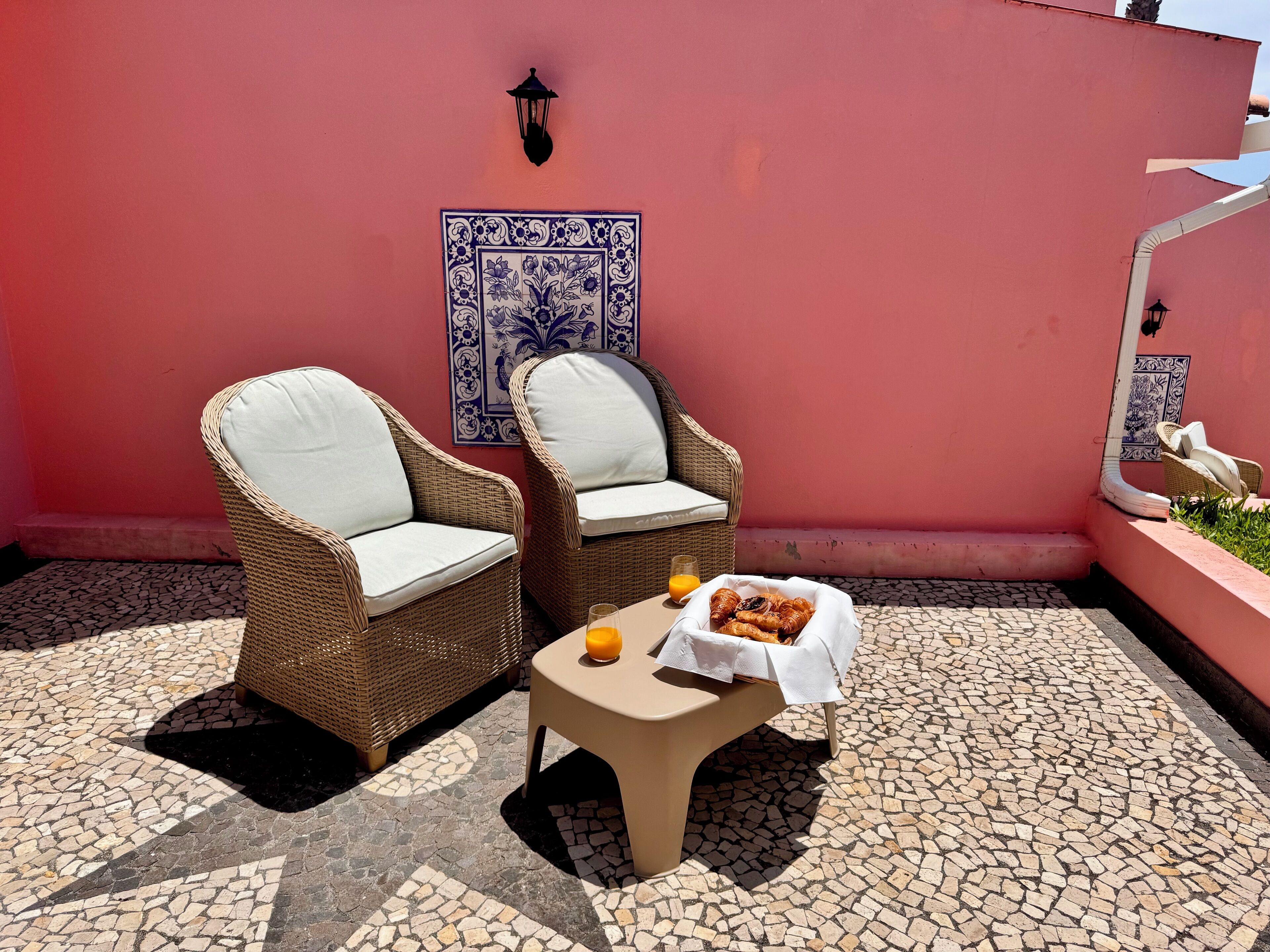Duas cadeiras de vime com almofadas brancas, uma mesa com uma cesta de pastelaria e um copo de sumo de laranja, e um chão em azulejos.
