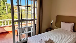 Hotelkamer met een bed, nachtkastje, lamp en uitzicht op een balkon met planten en een balustrade.