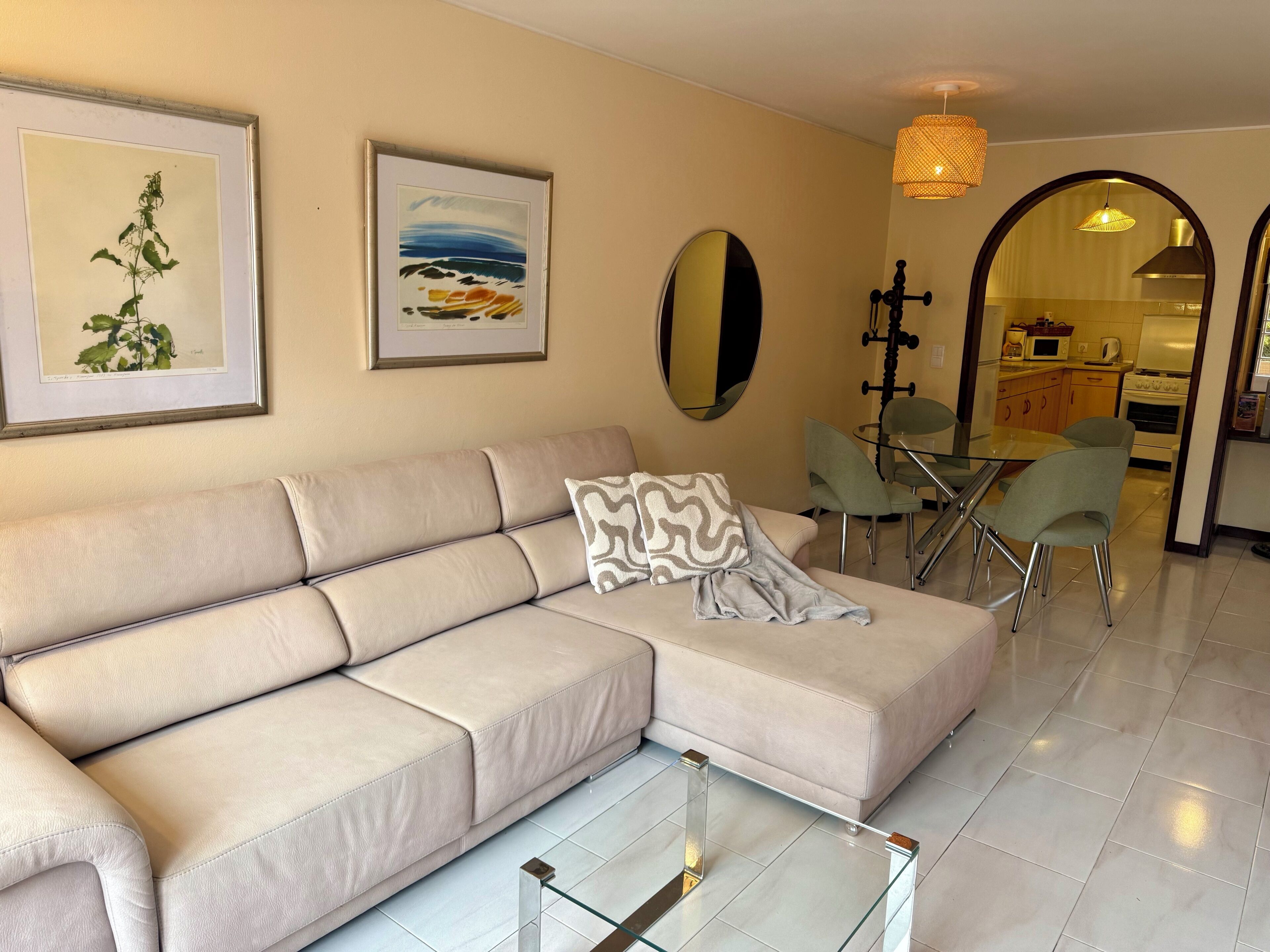 A living room with a beige sofa, a glass coffee table, a dining area with a glass table and chairs, and a kitchen visible through an archway.