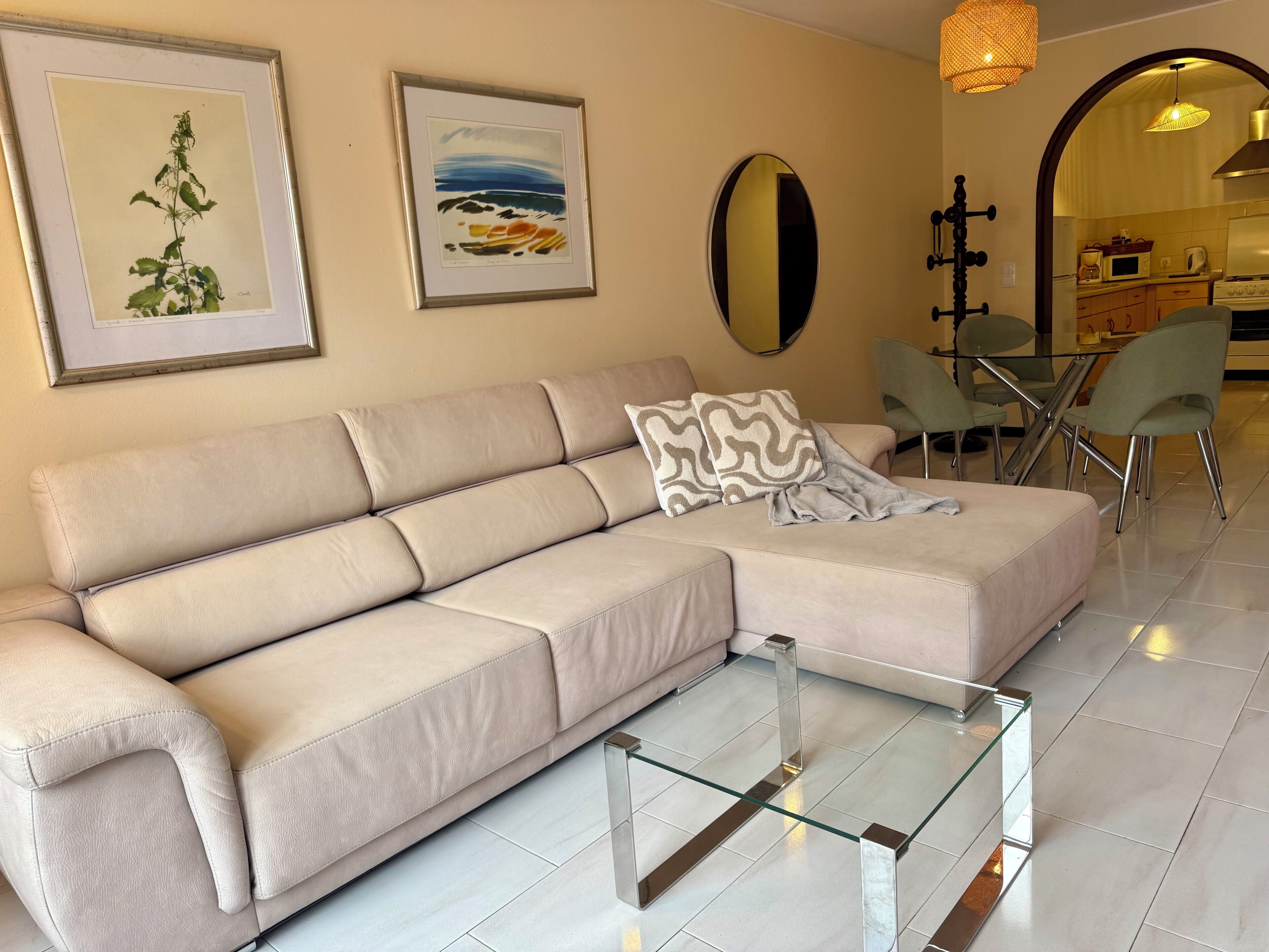 Un sofá beige con una mesa de centro de vidrio, impresiones botánicas enmarcadas en la pared y una cocina visible a través de un arco.