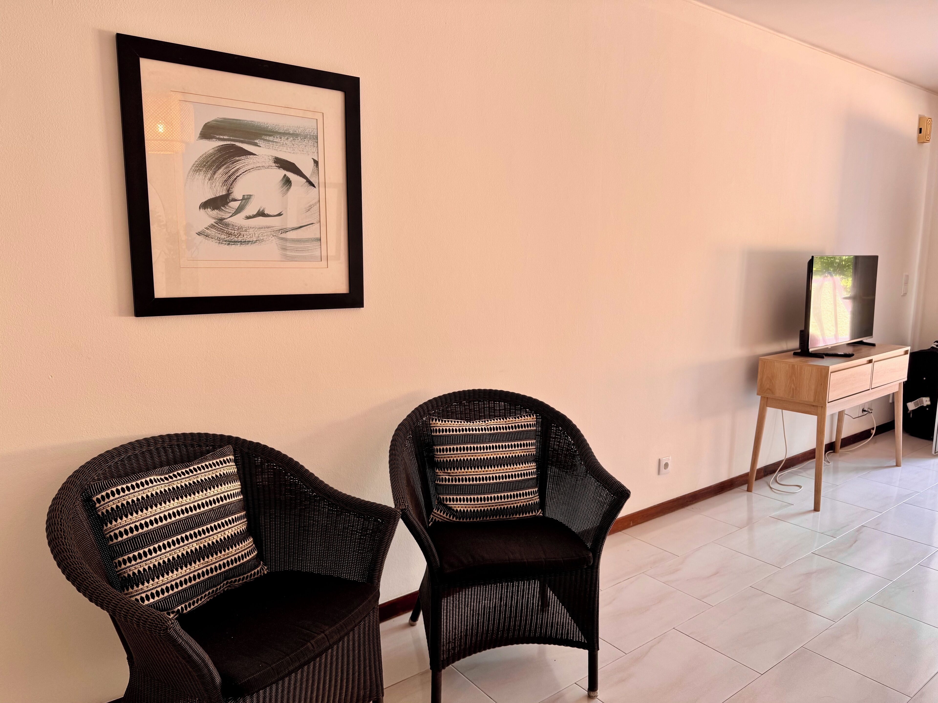 A room with two wicker chairs, a framed artwork on the wall, and a TV on a wooden stand.