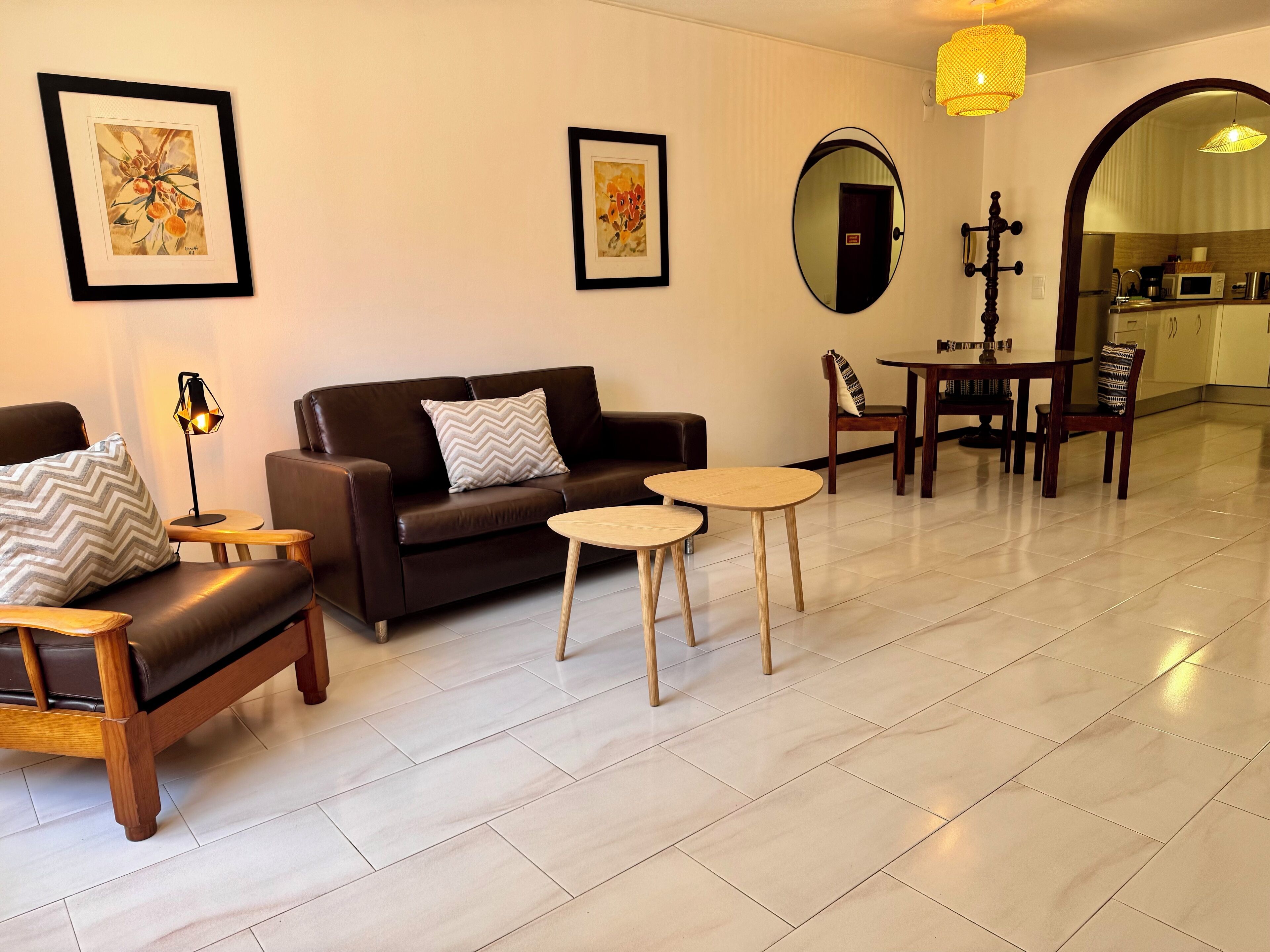 A modern living room with a brown leather sofa, two small round tables, and a wooden chair. There are framed artworks on the walls and a mirror above the sofa.