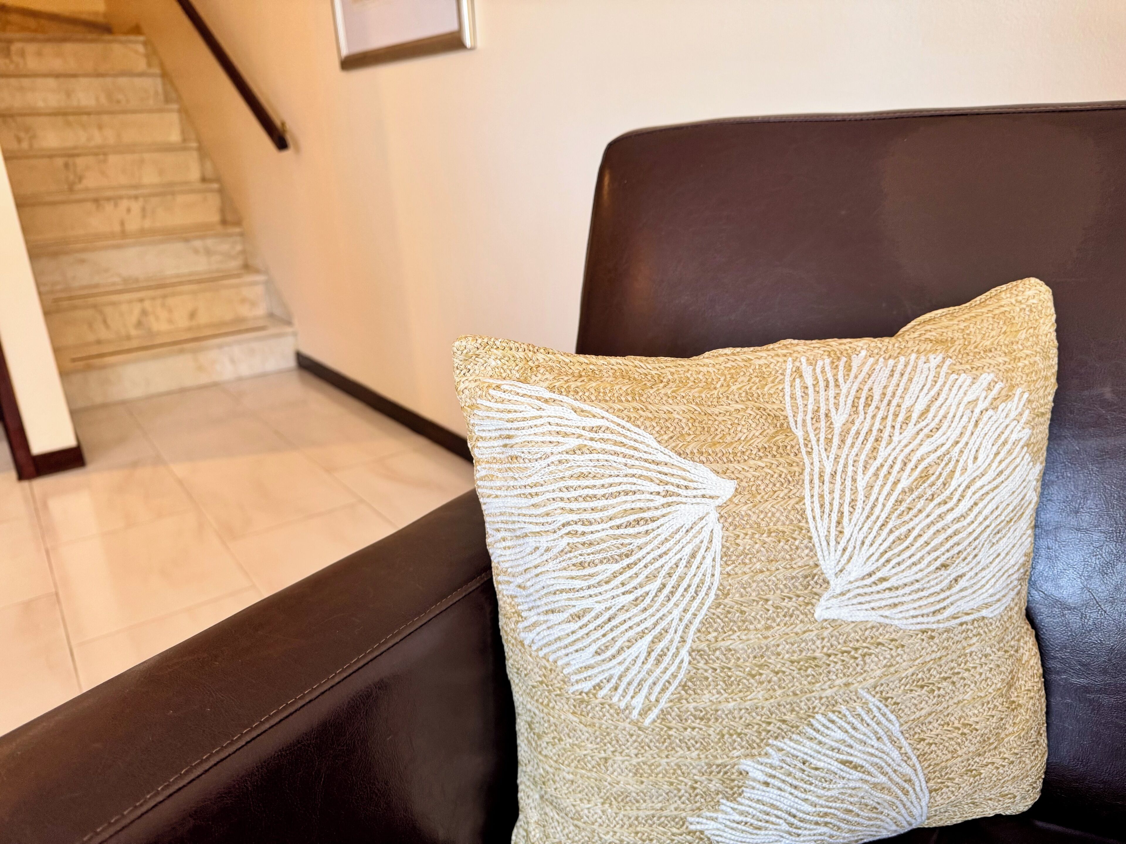 Un coussin beige à texture particulière, orné de motifs de feuilles blanches, posé sur un canapé en cuir foncé.