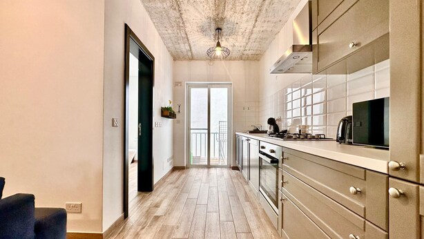 A modern kitchen with a wooden floor, stainless steel cabinets, and a large window. - Stylish Apt Off The Sea Front (Il-Gzira)