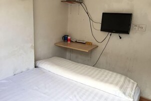 Una habitación pequeña con cama individual, televisión instalada en la pared, un estante con objetos y un enchufe.