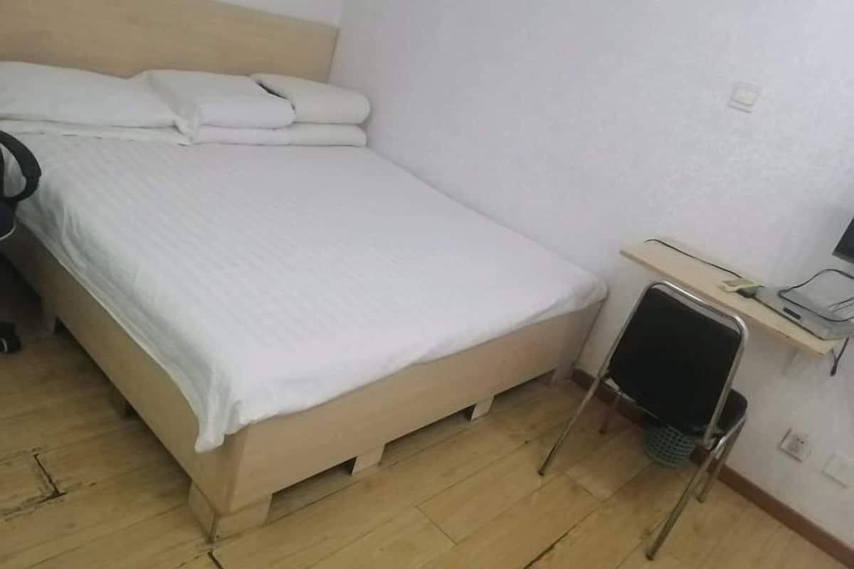 A small, neatly made bed with white linens, a wooden headboard, and a small desk with a chair beside it.