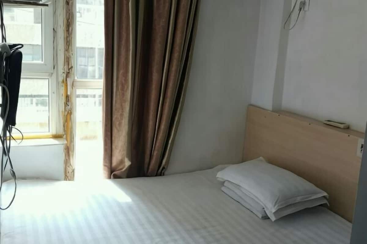 A single bed with a wooden headboard, a window with curtains, and a visible cable on the floor.