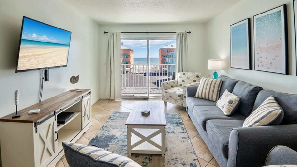 A modern living room with a large TV, a sofa, a coffee table, and a view of the beach through the balcony doors.