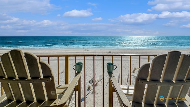 A beach view from a wooden deck with two Adirondack chairs and two teal mugs on the railing.