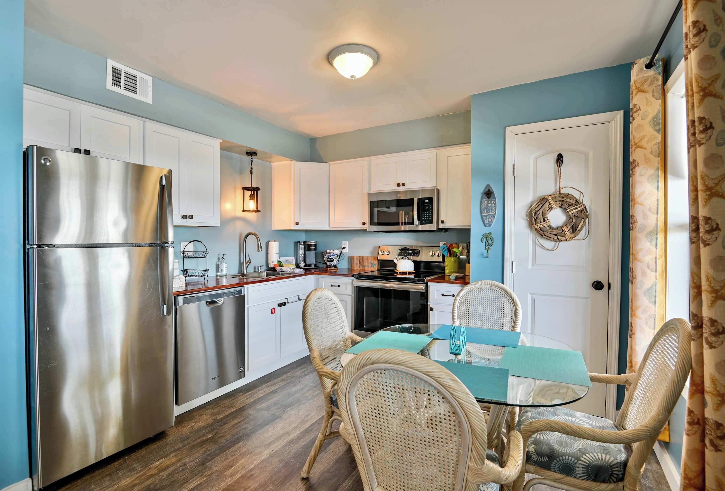 A compact kitchen with white cabinets, stainless steel appliances, wicker chairs, and a glass table.