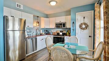 A compact kitchen with white cabinets, stainless steel appliances, wicker chairs, and a glass table.