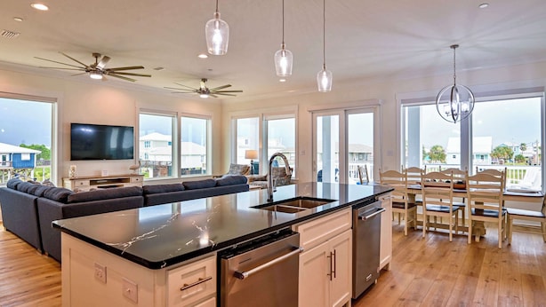 A spacious kitchen with a large island, white cabinetry, and stainless steel appliances. There is a dining area with wooden chairs and a large window offering a view of the outdoors.