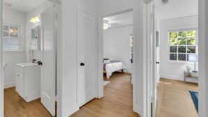 Un pasillo con puertas blancas que dan a un baño y un dormitorio. El baño tiene un lavamanos y un espejo. El dormitorio tiene una cama y una ventana.
