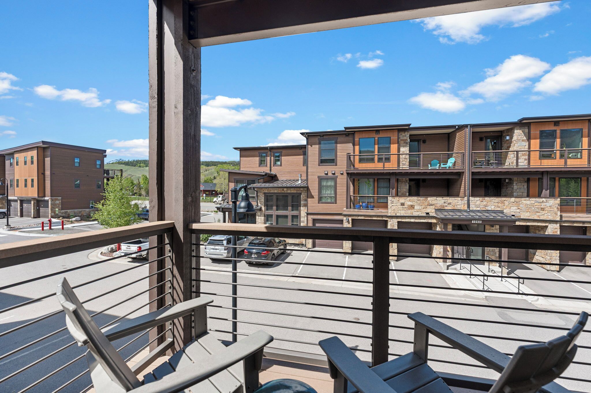 A balcony with chairs, a view of a multi-story building with balconies, and a parking area.