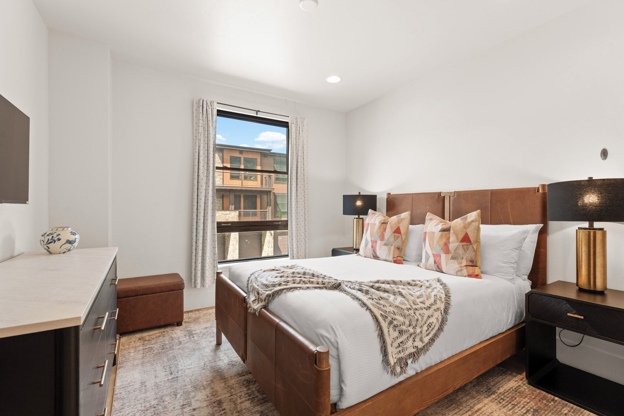 A modern hotel room with a bed, bedside tables, a desk, and a window with a view of buildings.