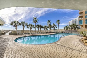 A swimming pool with lounge chairs and palm trees, surrounded by a brick patio and a building with balconies.
