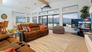 A spacious living room with a brown leather sofa, a flat-screen TV, and a large window offering a mountain view.