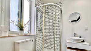 Une salle de bain moderne avec une douche à parois de verre, des murs recouverts de carreaux blancs, un miroir rond et un lavabo blanc.