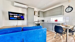 Une cuisine moderne avec des meubles gris, un four encastré et un four à micro-ondes. On y trouve une table à manger avec des chaises et un canapé bleu. Un téléviseur à écran plat est fixé au mur et diffuse différents services de streaming.