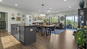 Une cuisine moderne dotée d’un îlot central, d’un coin repas avec une table et des chaises en bois, et d’un salon avec un canapé et une grande fenêtre.
