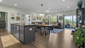 Une cuisine moderne dotée d’un îlot central, d’un coin repas avec une table et des chaises en bois, et d’un salon avec un canapé et une grande fenêtre.