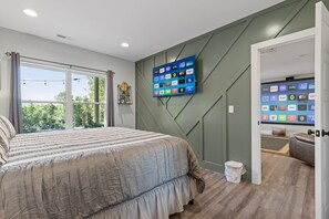 Un dormitorio con una cama grande, un televisor montado en la pared mostrando diversas plataformas de streaming, y una ventana con cortinas.