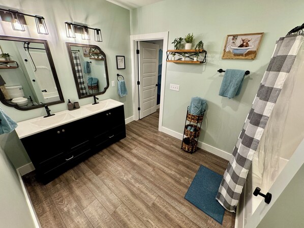 Un baño con tocador, espejos, cortina de ducha, alfombra azul y una puerta que da a otra habitación.