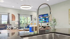A modern kitchen with a stainless steel sink, a granite countertop, and a flat-screen TV mounted on the wall.