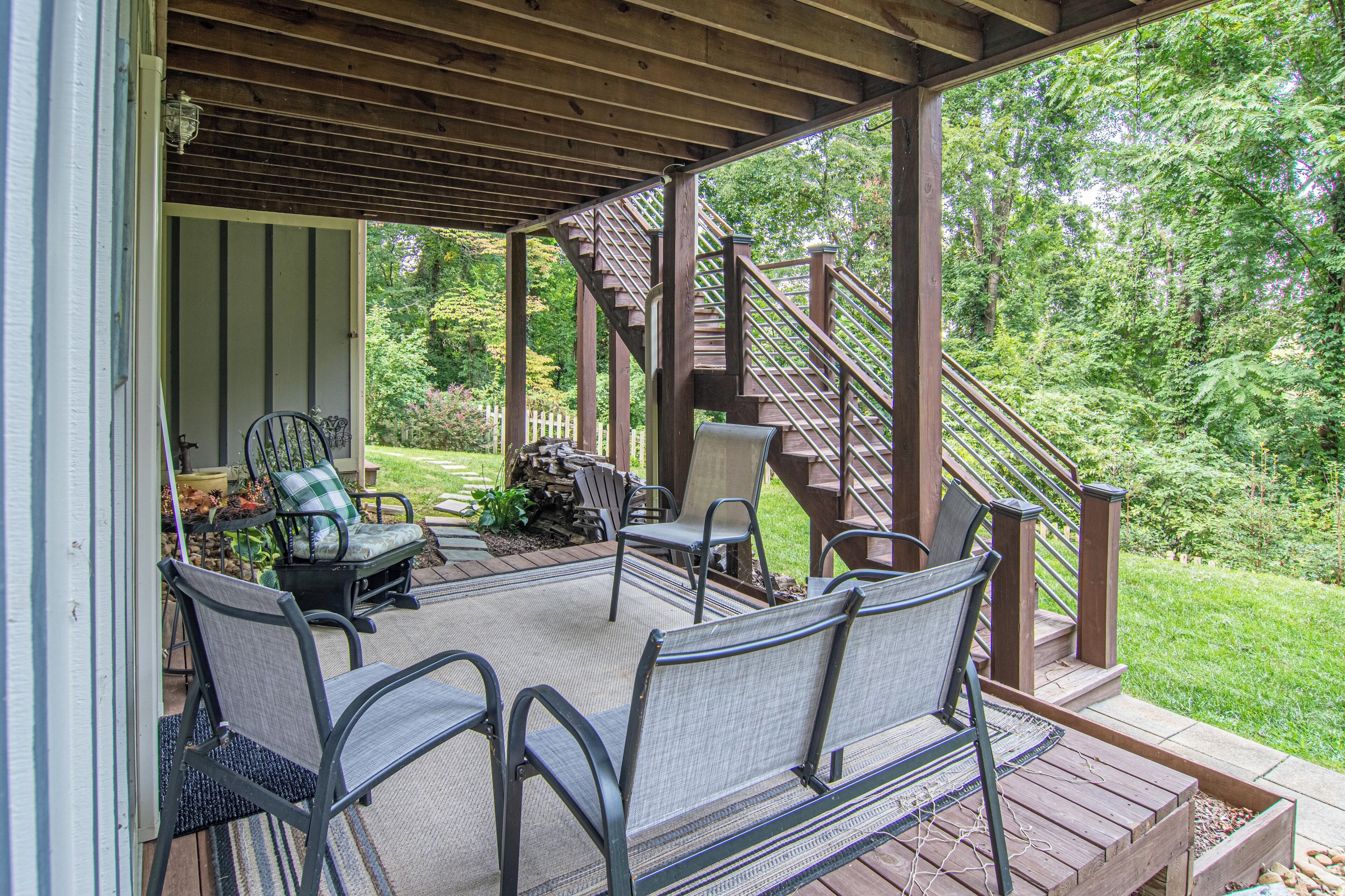 Eine überdachte Terrasse im Freien mit einer Holztreppe, einer Sitzecke mit Stühlen und Blick auf einen üppigen Garten.