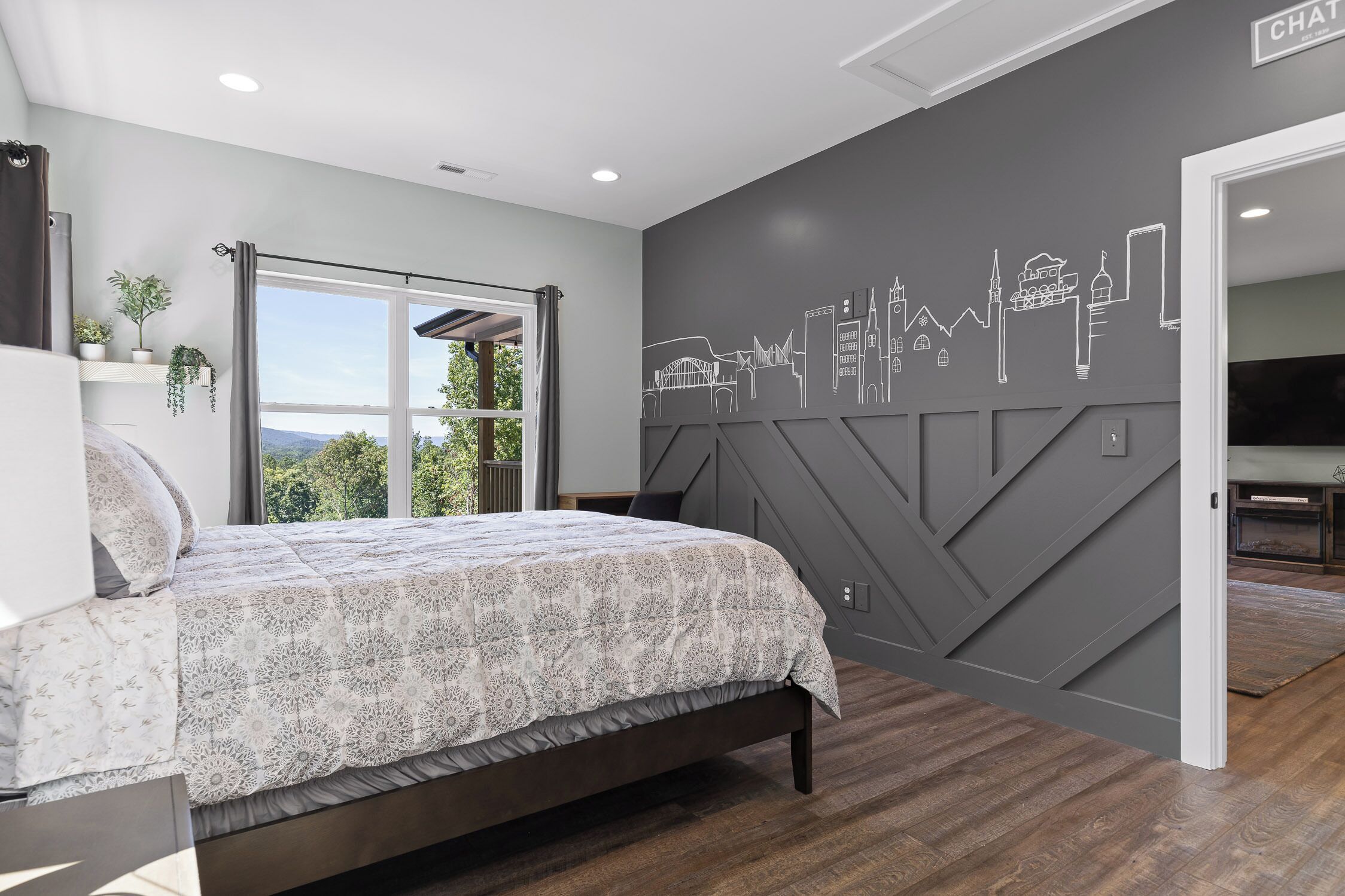 A bedroom with a bed, a cityscape wall mural, a window with curtains, and a TV in the adjacent room.