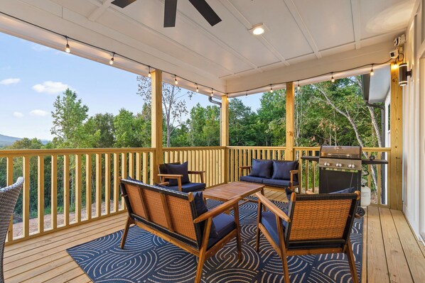 A covered outdoor deck with wooden furniture, a grill, and a view of the surrounding greenery.