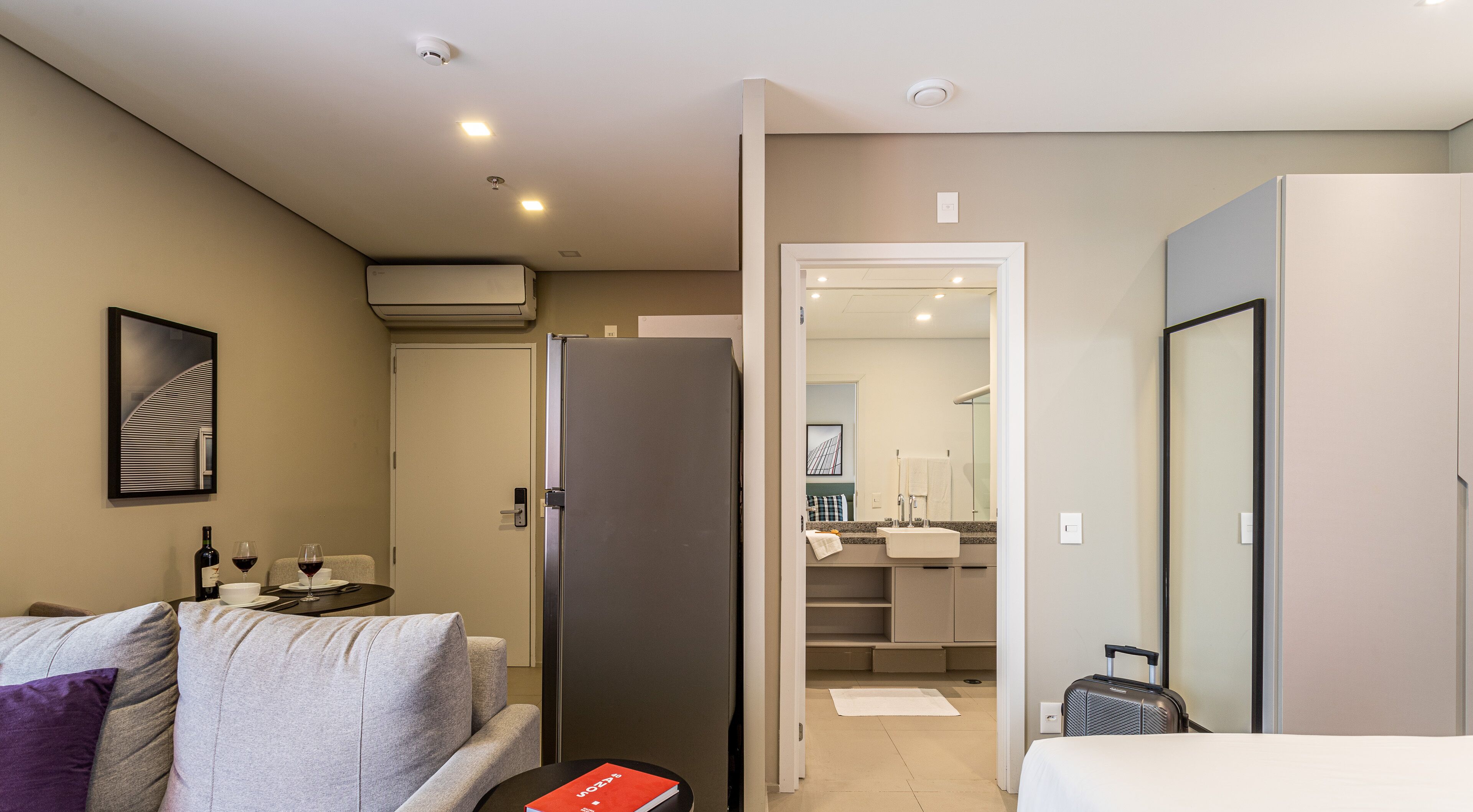 A hotel room with a bed, a sofa, a dining table with wine glasses, a refrigerator, a bathroom visible through an open door, and a suitcase on the floor.