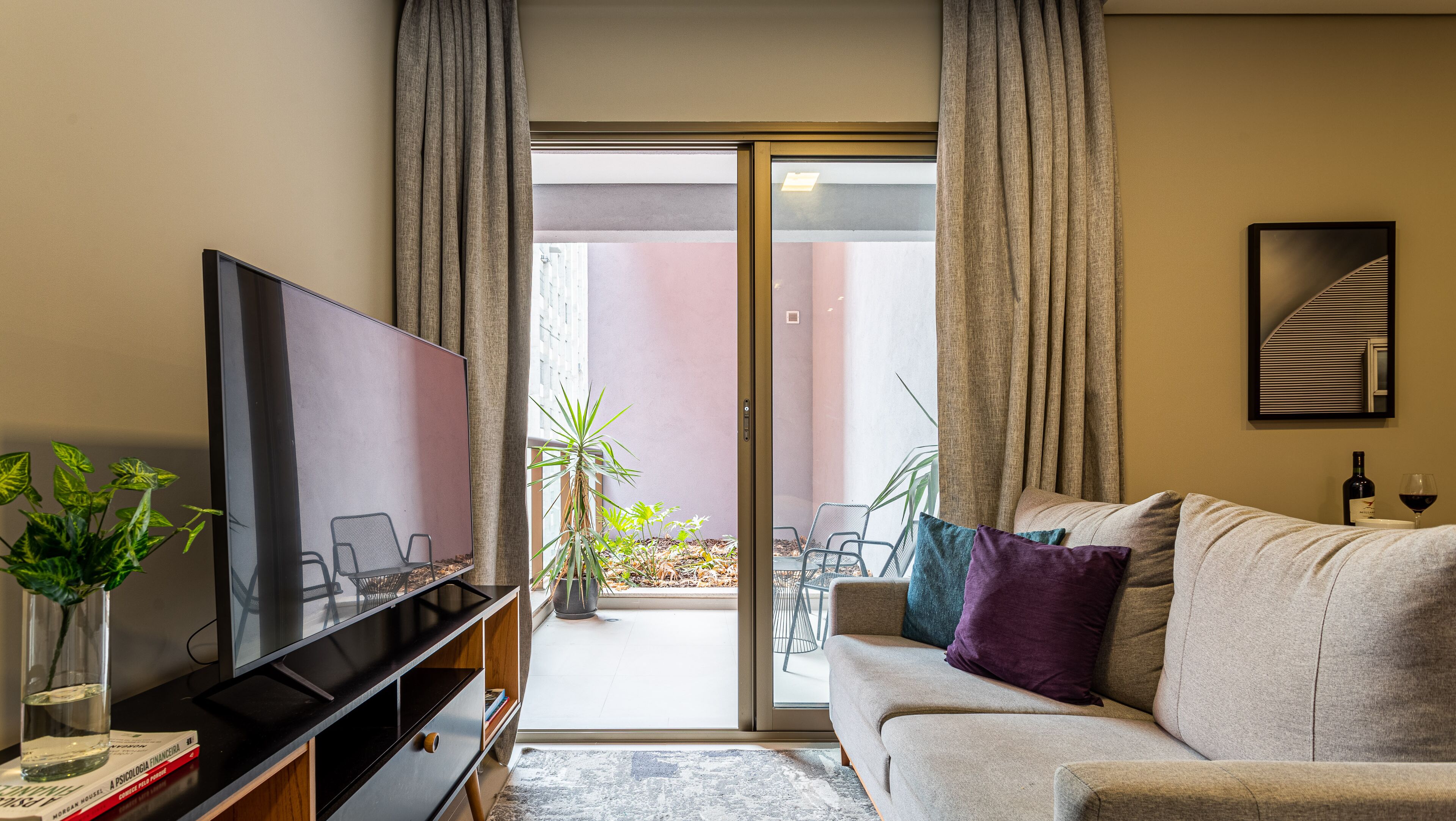 A living room with a TV, sofa, and a sliding glass door leading to an outdoor area with plants.