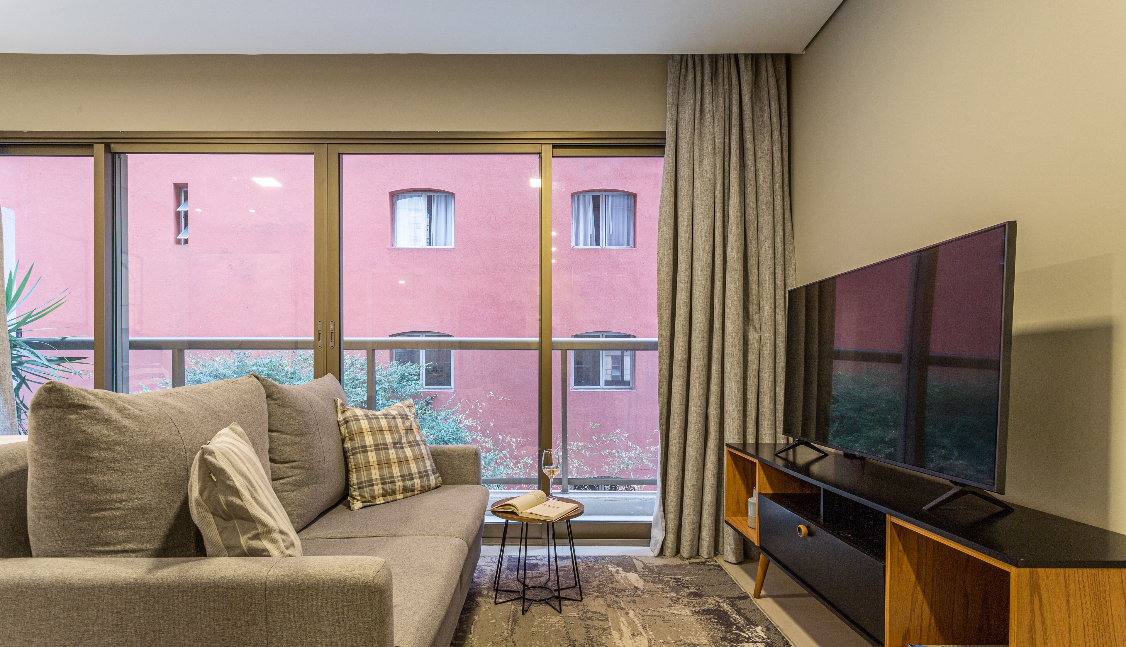 A living room with a sofa, a TV, and a view of a red building through large windows.